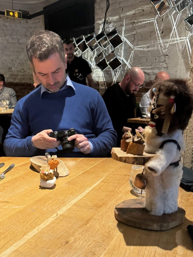 Άνδρας σε εστιατόριο που φωτογραφίζει φαγητό, με μια γλυκιά δημιουργία σε ένα ξύλινο πιάτο. Στο πλάι, μια φιγούρα σε στυλ κουζίνας κρατάει ακόμα ένα πιάτο.